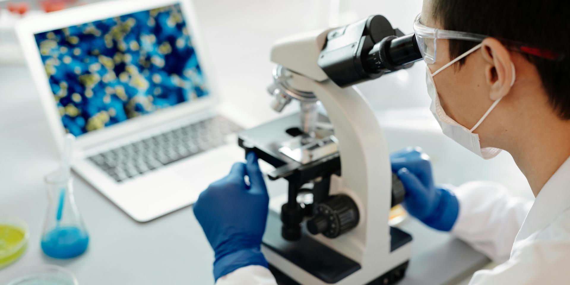 Person wearing white coat looking into microscope, with laptop in background