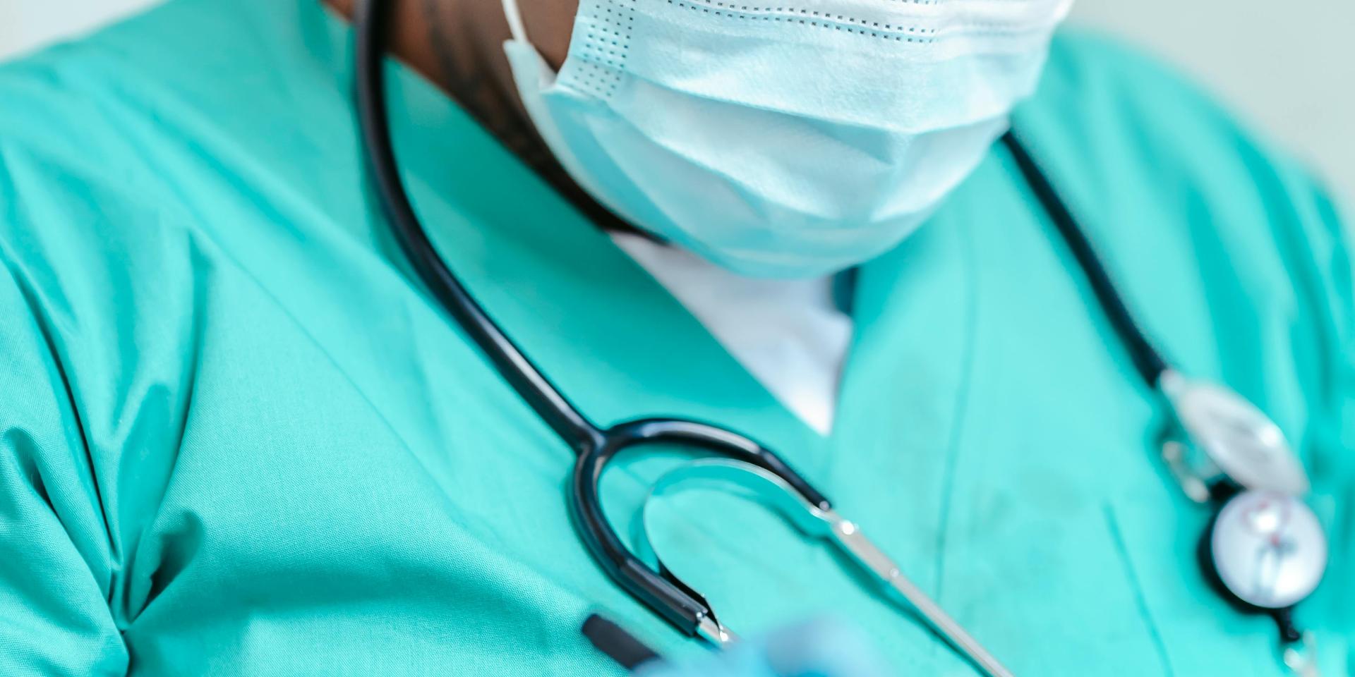 Person wearing mask, stethoscope, green scrubs and blue gloves writing on clipboard