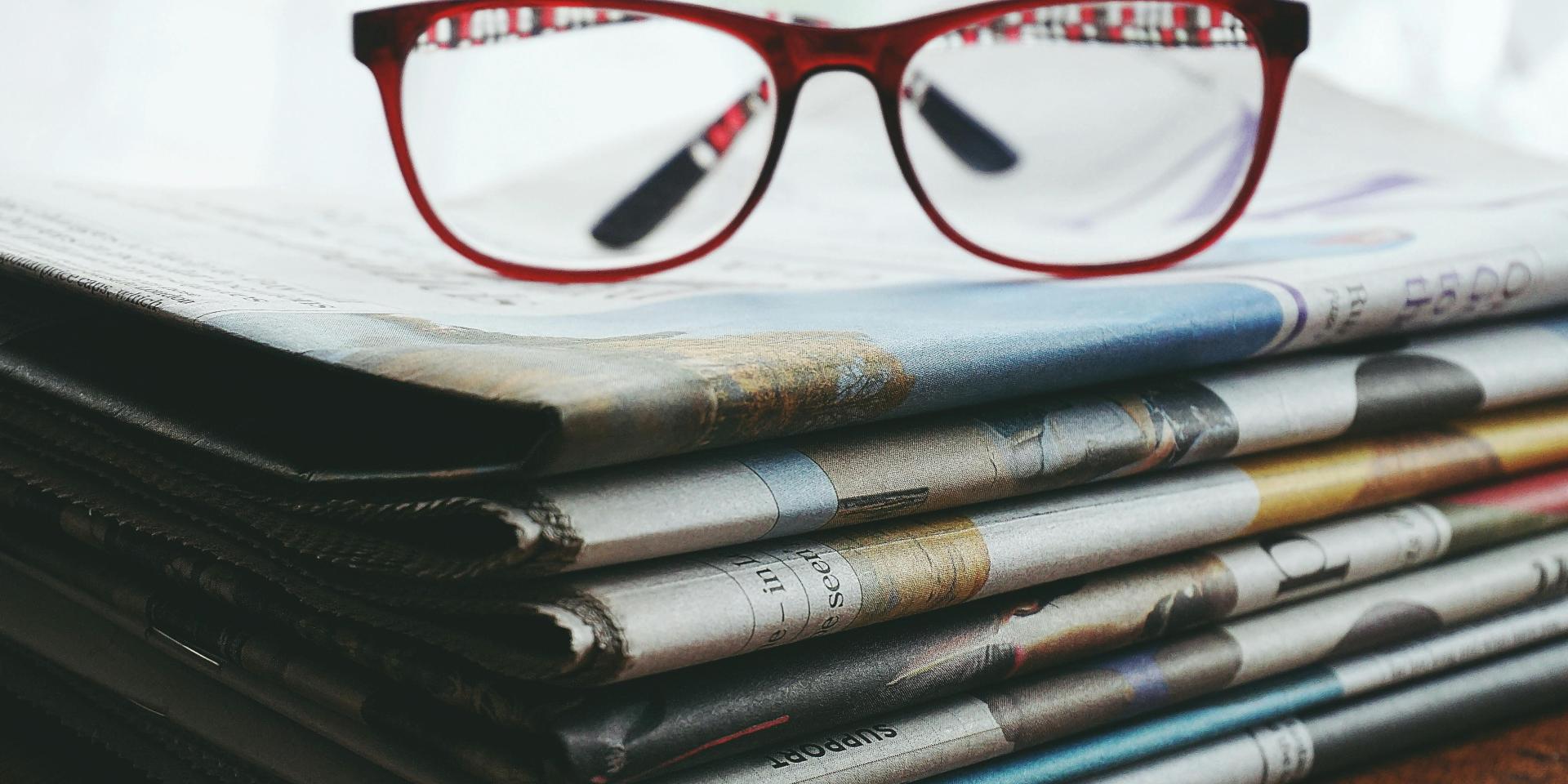 A pair of red framed eye glasses sitting on top of a stack of newspapers