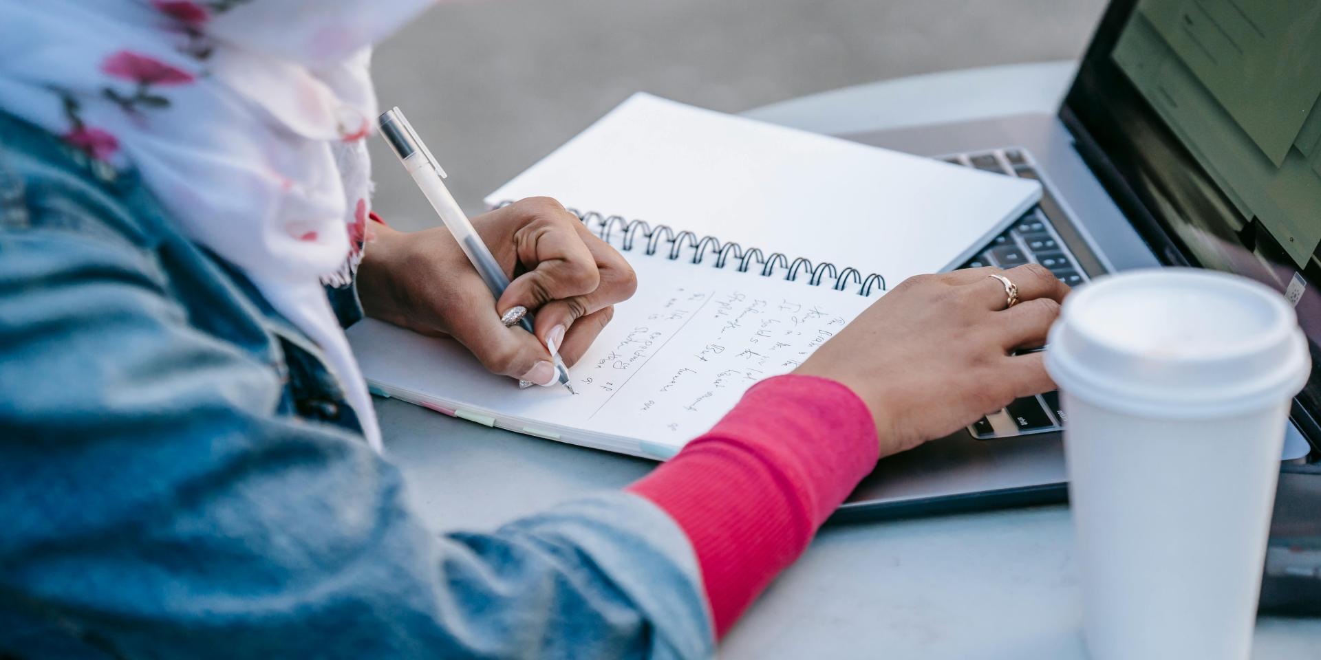 A person sits at a table outdoors, writing in a notebook and typing on a laptop