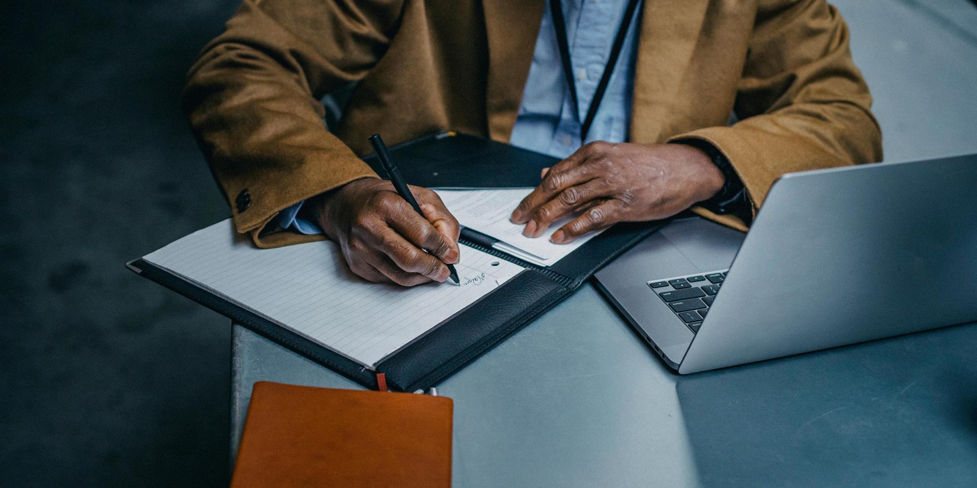 A person sitting at a table uses a laptop and writes in a notebook