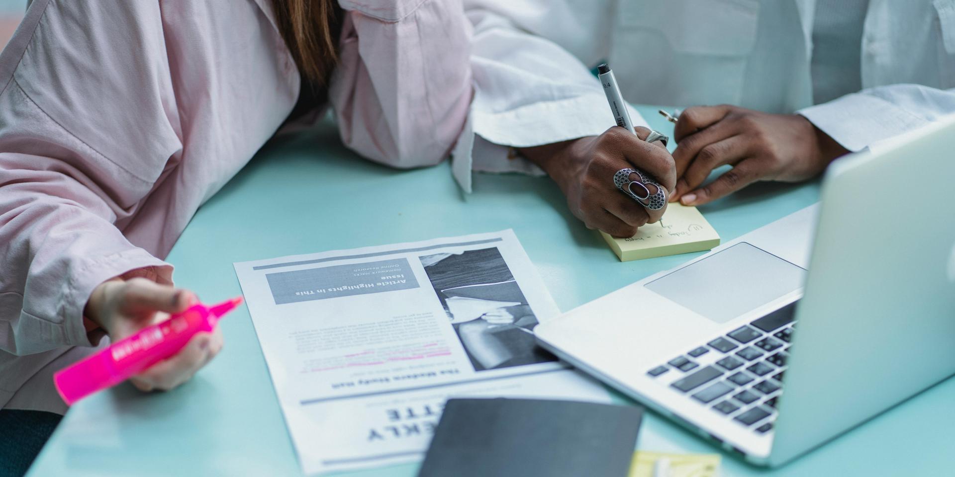 People collaborating at a table with a laptop, documents, notebook, sticky notes and writing implements