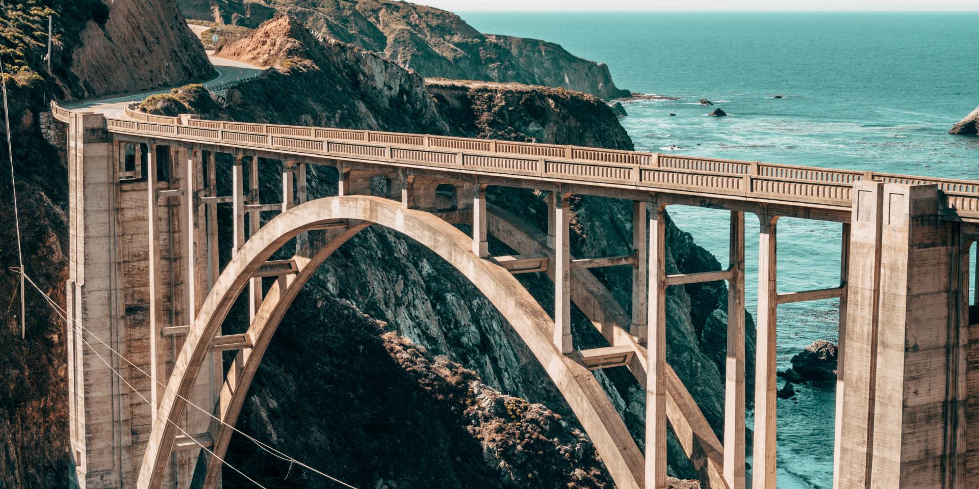 Tan bridge between a rocky cliff above teal water