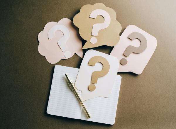 Paper cut outs of think bubbles with question marks in the middle, next to a writing implement and notebook