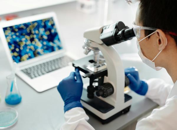 Person wearing white coat looking into microscope, with laptop in background