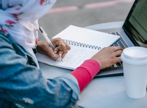 A person sits at a table outdoors, writing in a notebook and typing on a laptop