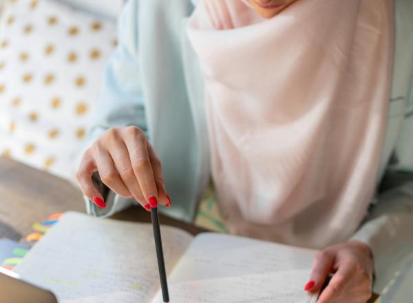 A person consults a notebook, pointing to its pages with a pencil