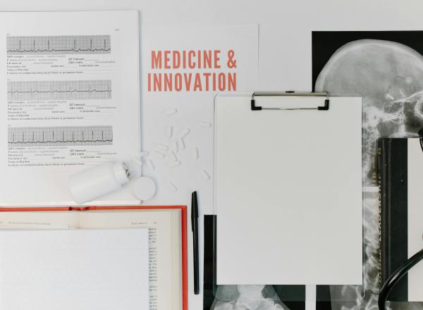 A white table with notebooks, textbooks, pill bottle spilling out pills, an x-ray, clipboard, a writing implement and a stethoscope