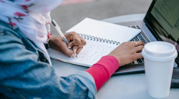 A person sits at a table outdoors, writing in a notebook and typing on a laptop